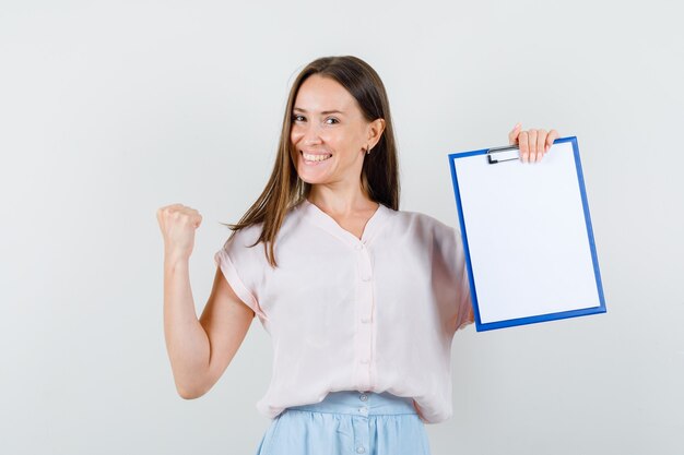 Young lady holding clipboard with winner gesture in t-shirt, skirt and looking joyful. front view.