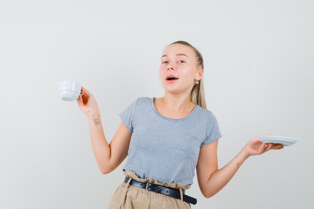 Giovane donna che tiene tazza di bevanda in maglietta, pantaloni e che sembra gioiosa. vista frontale.