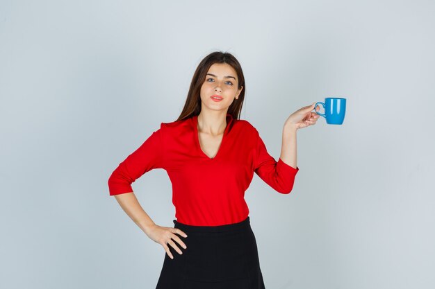 Young lady holding cup while keeping hand on hip in red blouse