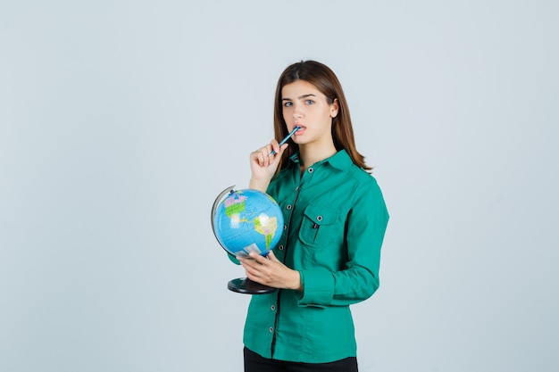 Young lady holding earth globe, keeping pen in mouth in shirt and looking thoughtful , front view.