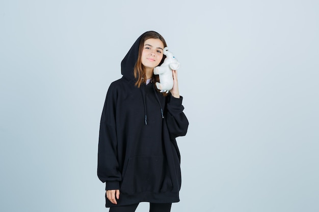 Young lady holding elephant toy on cheek in oversized hoodie, pants and looking pretty , front view.