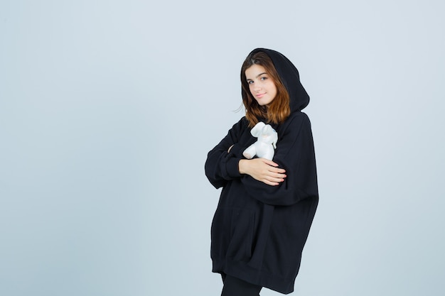 Free photo young lady holding elephant toy on chest in oversized hoodie, pants and looking cute , front view.