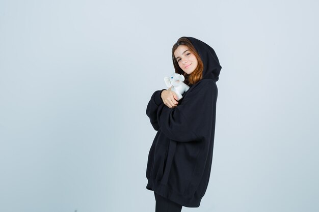 Young lady holding elephant toy on chest in oversized hoodie, pants and looking pretty , front view.