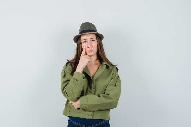 Young lady holding finger on cheek in jacket pants hat and looking upset 