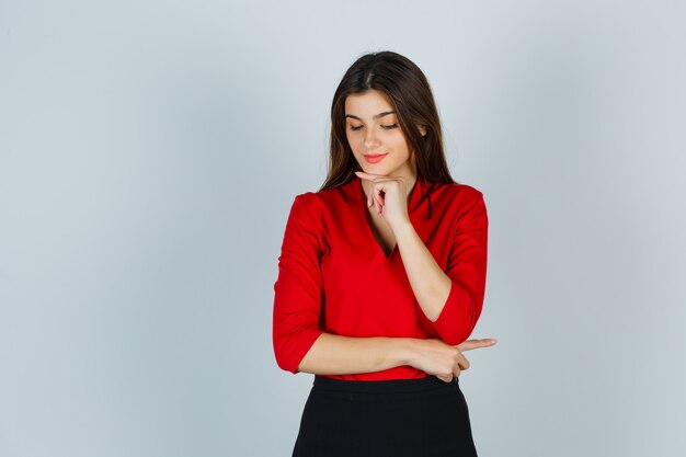 Young lady holding finger under chin in red blouse, skirt and looking gorgeous