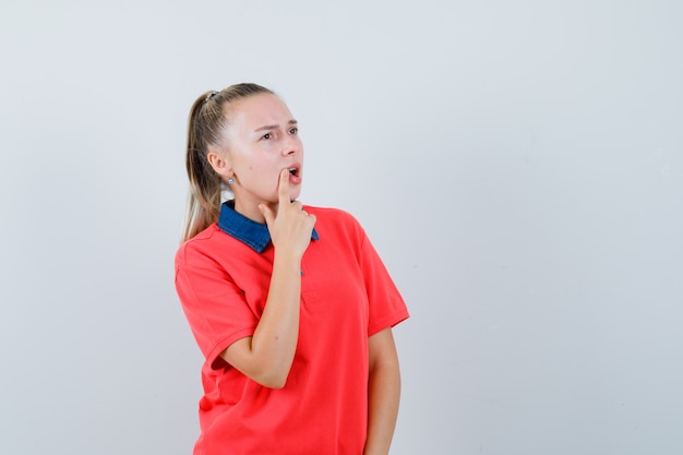 Young lady holding finger near mouth in t-shirt and looking pensive. front view.