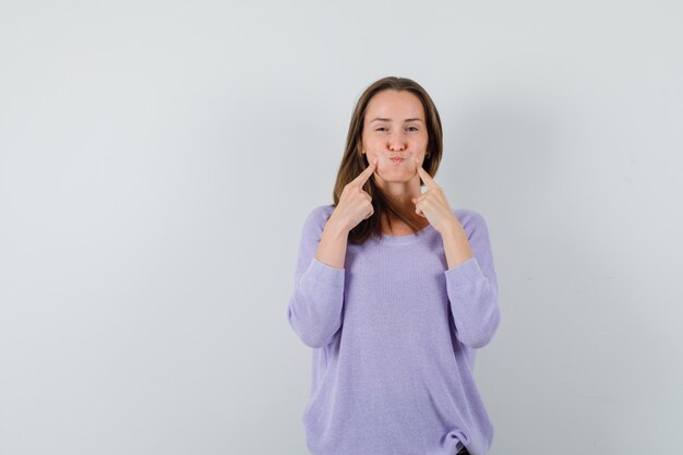 Young lady holding fingers on blown cheeks in casual shirt and looking funny. front view.