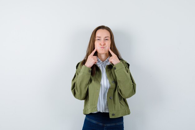 Young lady holding fingers on blown cheeks in shirt, jacket and looking lovely. front view.