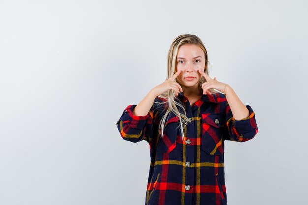 Young lady holding fingers on cheeks in checked shirt and looking cute , front view.