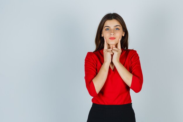 Young lady holding fingers on cheeks in red blouse, skirt and looking pretty