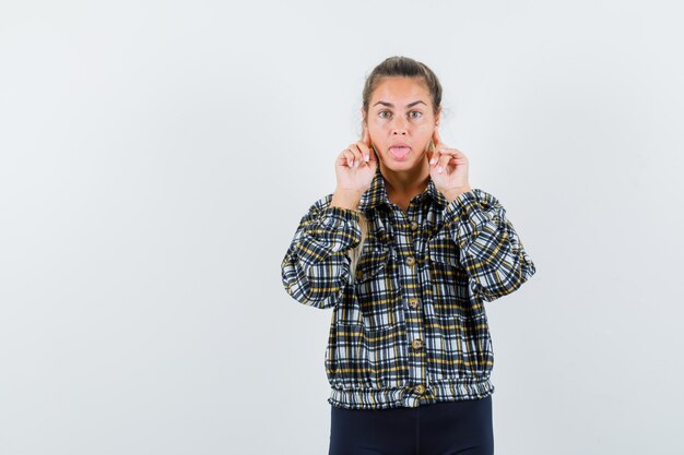 Young lady holding fingers on cheeks, sticking out tongue in shirt, shorts and looking pensive , front view.