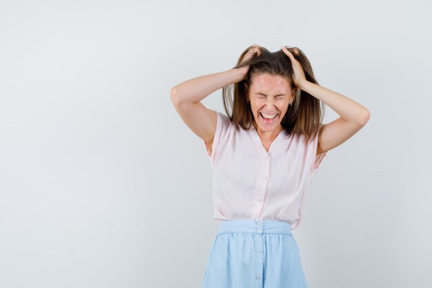 Young lady holding fingers in hair in t-shirt, skirt and looking blissful. front view.