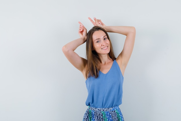 Young lady holding fingers over head as bull horns in singlet, skirt and looking funny