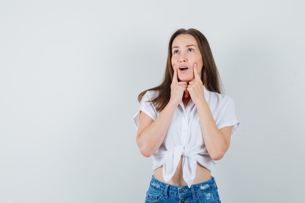 Young lady holding fingers on her cheeks in white blouse and looking funny. front view. space for text