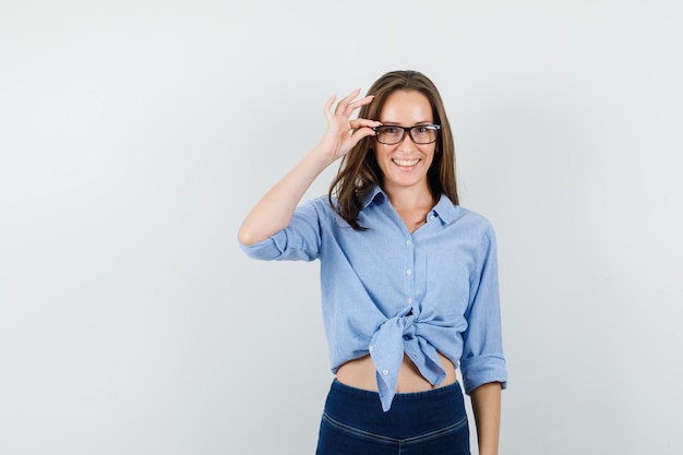 Young lady holding fingers on her glasses in blue shirt, pants and looking optimistic