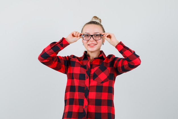 Young lady holding fingers on her glasses in checked shirt and looking jolly. front view.