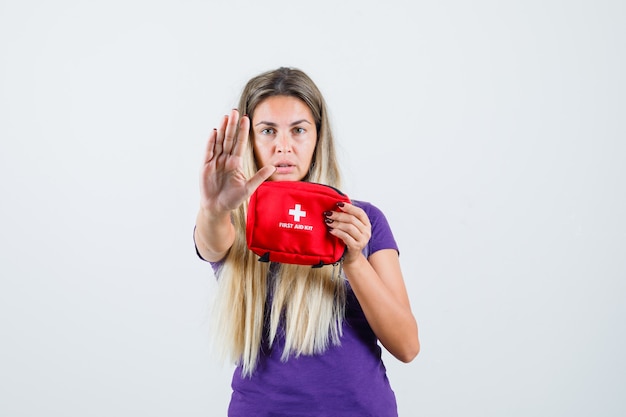 Foto gratuita giovane donna che tiene il kit di pronto soccorso, mostrando il gesto di arresto in maglietta viola, vista frontale.