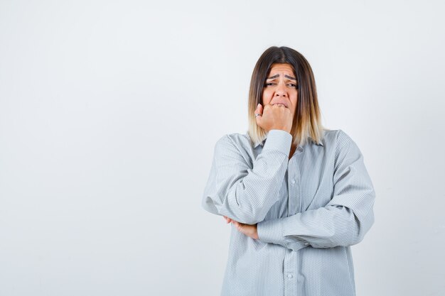 Young lady holding fist on mouth in oversized shirt and looking scared , front view.