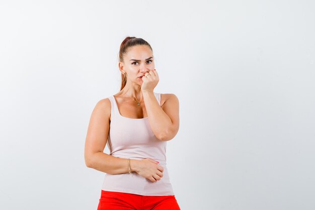 Young lady holding fist near mouth in beige tank top and looking careful