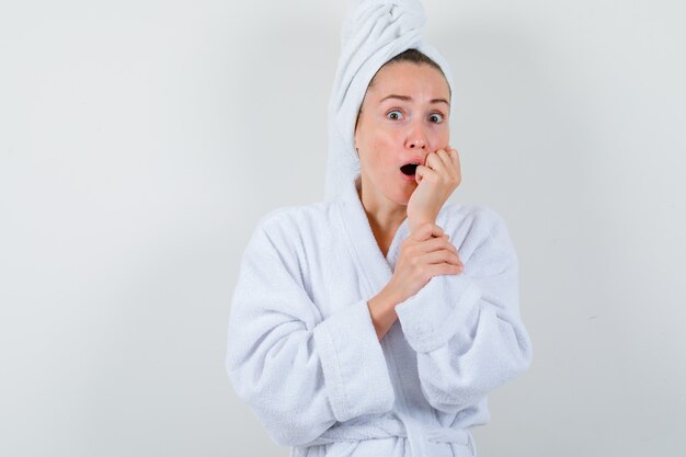 Young lady holding fist near mouth in white bathrobe, towel and looking surprised , front view.