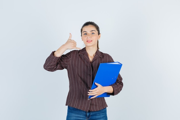 Young lady holding folder, showing phone gesture in shirt, jeans and looking wistful, front view.