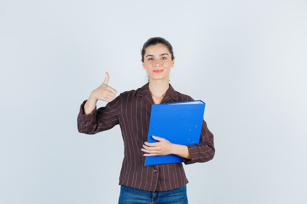 Young lady holding folder, showing thumb up in shirt, jeans and looking pleased , front view.