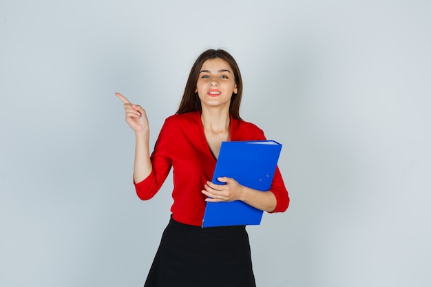 Young lady holding folder while pointing at upper left corner in red blouse