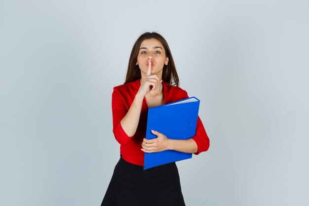 Young lady holding folder while showing silence gesture in red blouse