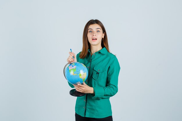 Young lady holding globe and pointing on it in shirt and looking perplexed , front view.