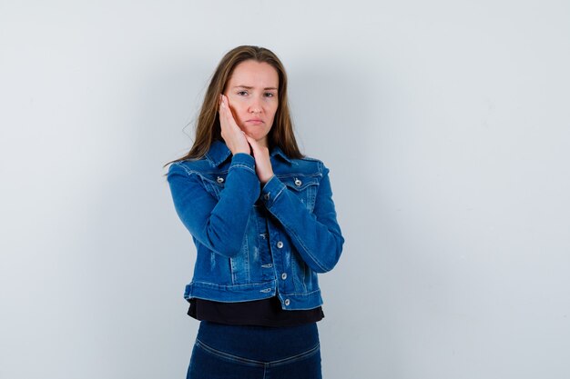 Young lady holding hand on cheek in blouse, jacket and looking sad , front view.