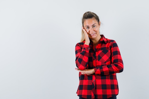 Young lady holding hand on cheek in checked shirt and looking dreamy