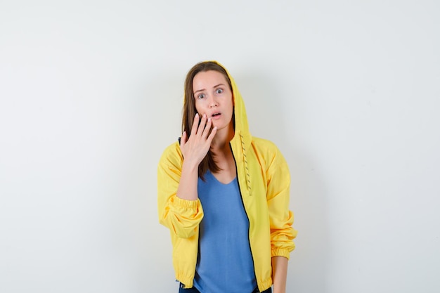 Young lady holding hand on cheek in t-shirt, jacket and looking surprised