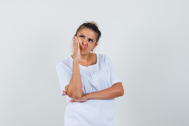 Young lady holding hand on cheek in t-shirt and looking pensive  