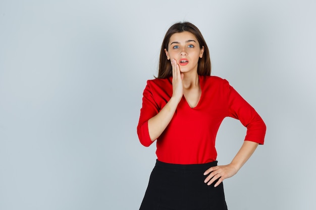 Young lady holding hand on cheek while keeping hand on hip in red blouse