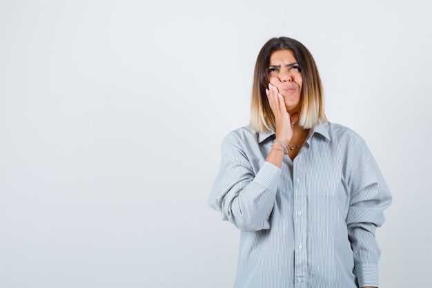 Young lady holding hand on cheek while looking up in oversized shirt and looking thoughtful , front view.