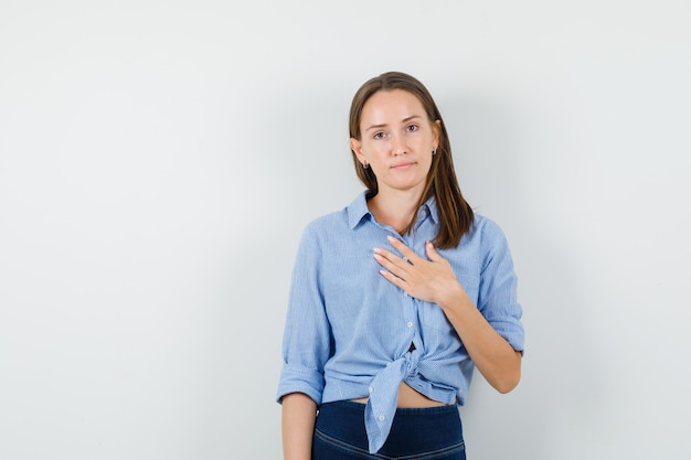 Giovane donna che tiene la mano sul petto in camicia blu, pantaloni e sguardo positivo.