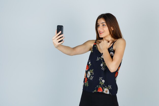 Young lady holding hand on chest while making video call in blouse and looking merry. front view.