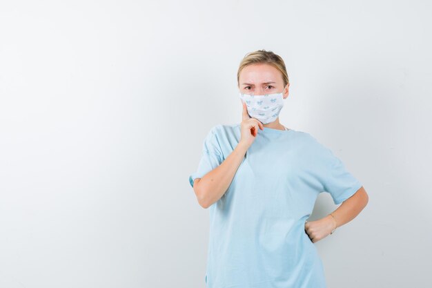 Young lady holding hand on chin in t-shirt, mask and looking serious