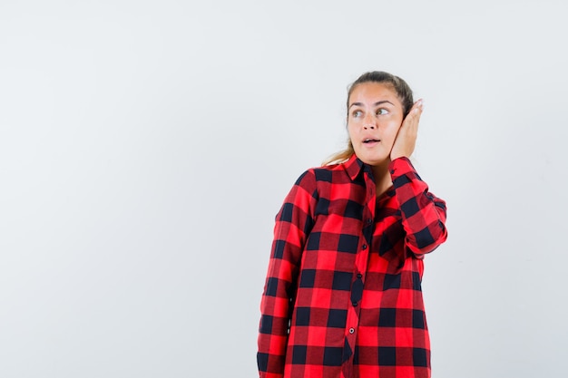 Young lady holding hand on ear in checked shirt and looking cute