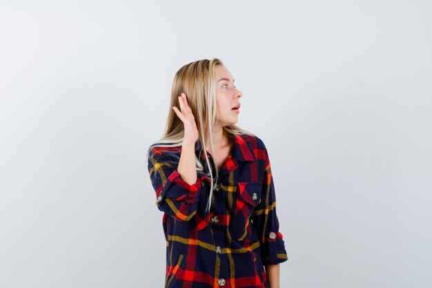 Young lady holding hand behind ear in checked shirt and looking wondered , front view.