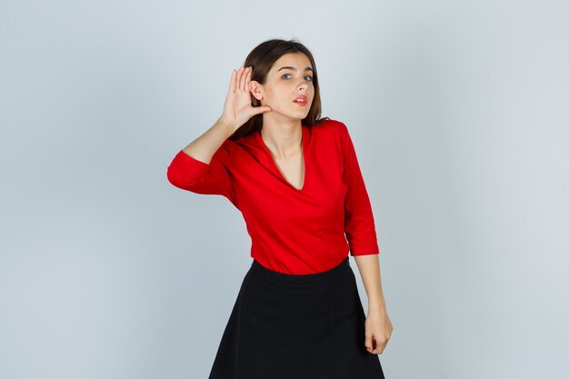 Young lady holding hand behind ear in red blouse, skirt and looking curious