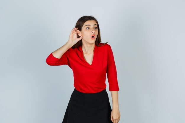 Young lady holding hand behind ear in red blouse, skirt and looking puzzled