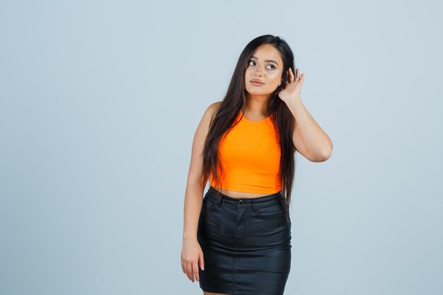 Young lady holding hand behind ear in singlet, mini skirt and looking focused. front view.