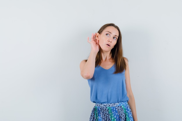 Young lady holding hand behind ear in singlet, skirt and looking curious