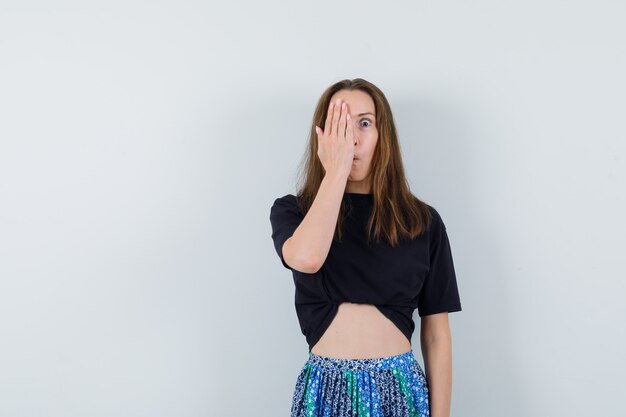 Young lady holding hand on eye in blouse, skirt and looking funny