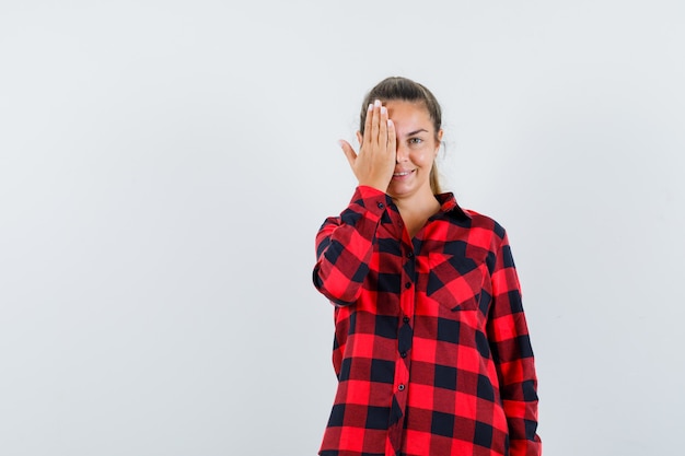 Young lady holding hand on eye in checked shirt and looking cheery