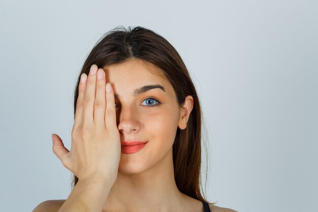 Young lady holding hand on eye and looking cheerful. front view.