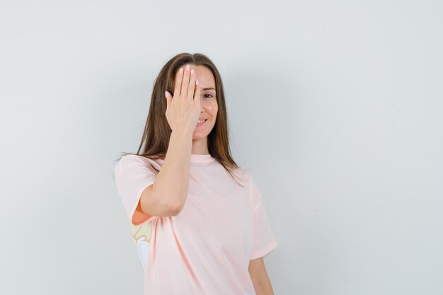 Young lady holding hand on eye in pink t-shirt and looking cheery  