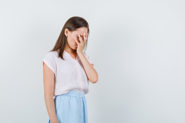 Young lady holding hand on face in t-shirt, skirt and looking pensive , front view.