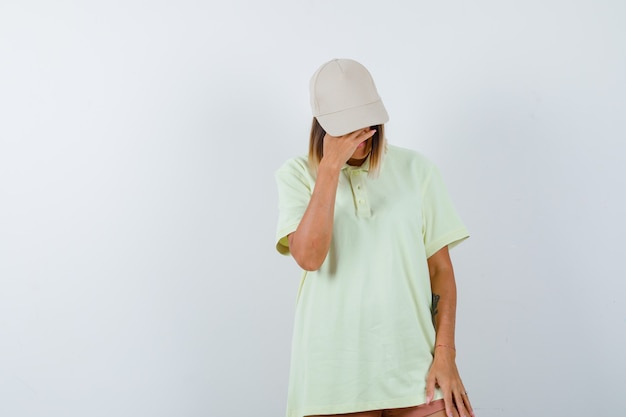 Young lady holding hand on forehead while looking down in t-shirt, cap and looking ashamed. front view.